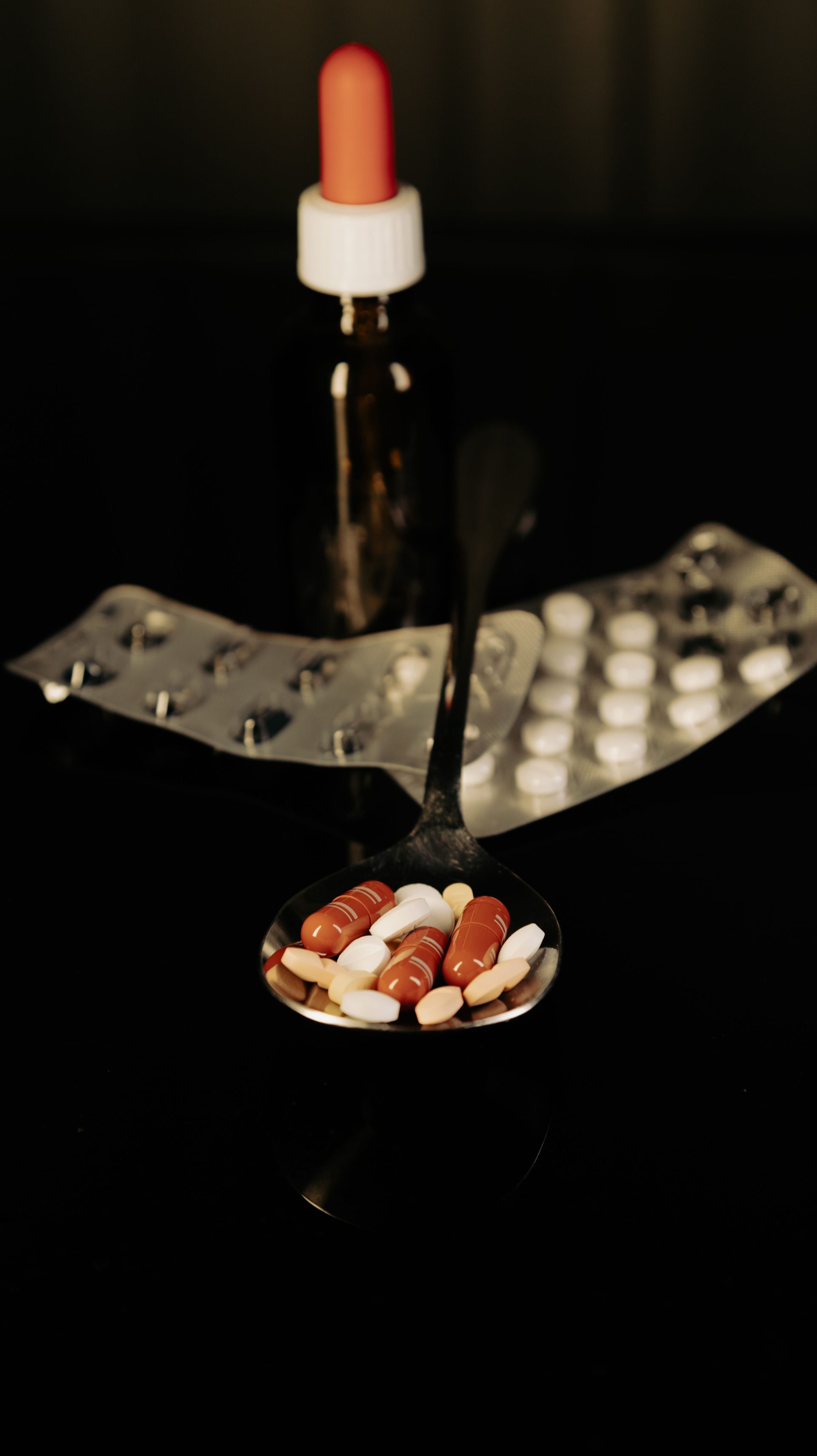 Nahaufnahme verschiedener Tabletten auf einem Löffel, mit Blisterpackungen und einer Pipette im Hintergrund.