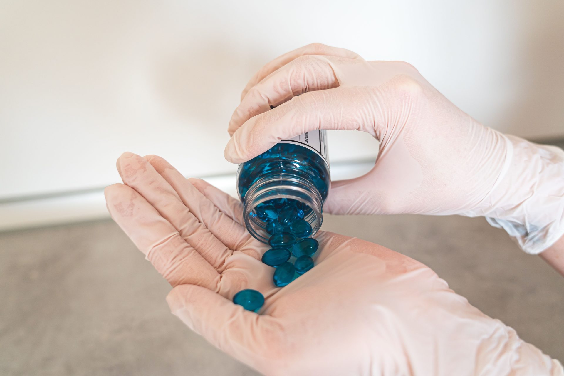 Nahaufnahme von behandschuhten Händen, die blaue Tabletten aus einer Flasche schütten – ein Symbol für Medikamente und Gesundheitsversorgung.